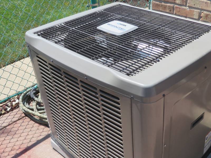 Outdoor condenser for a central air conditioning system on a concrete pad beside a brick wall, with a chain-link fence and a coiled garden hose