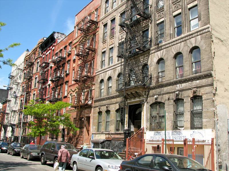 Row of prewar Manhattan apartment buildings with black fire escapes zigzagging down their brick and stone fronts, parked cars and a street tree