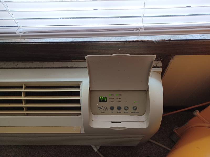 Packaged terminal air conditioner under an apartment window, its control panel cover open and the display reading 74