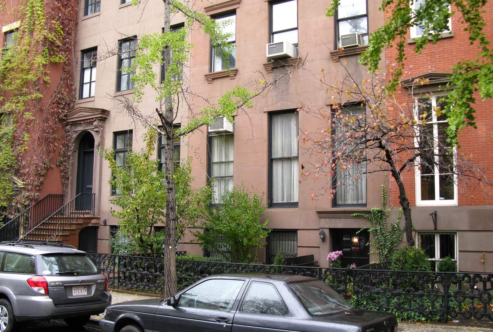 Brownstone and brick row houses on St. Luke's Place in Greenwich Village, Manhattan, with window air conditioners in several upper-floor windows, street trees, an iron fence and cars parked at the curb