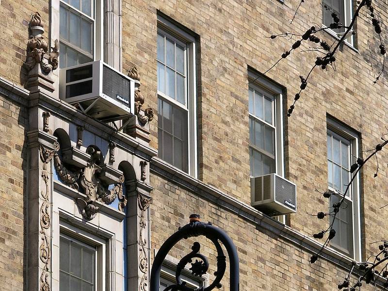 Two window air conditioners in the windows of a tan brick prewar apartment building on West 9th Street, with carved stone trim and the top of a cast-iron lamppost