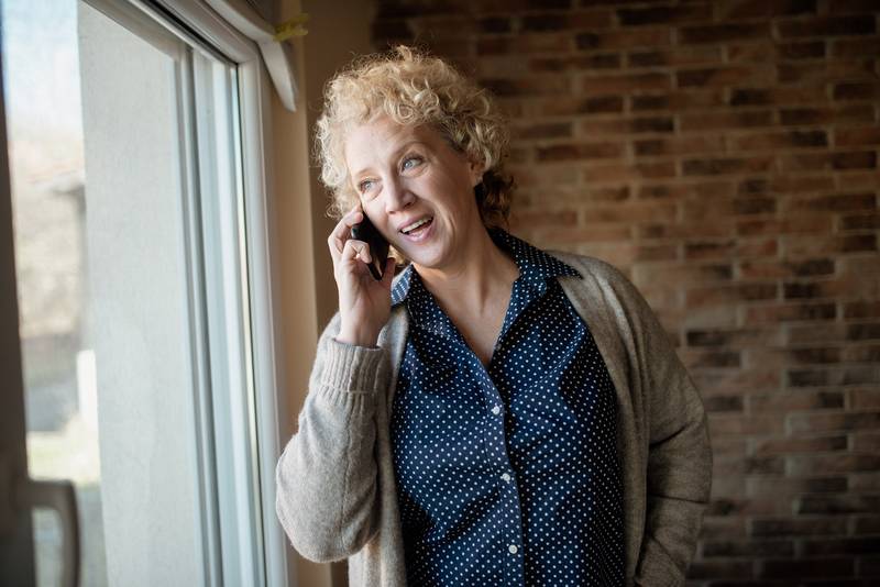A woman with curly hair smiling while she talks on a cell phone beside a window in a room with an exposed brick wall
