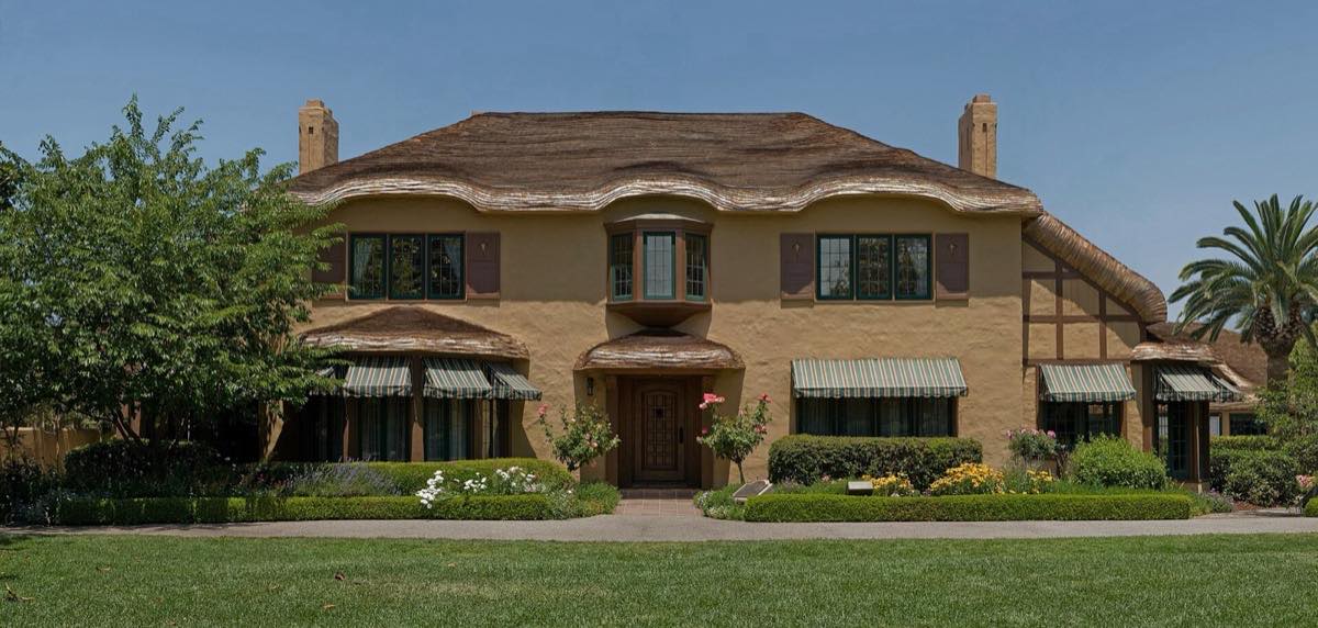 The Ainsley House, a 1925 English Tudor cottage with a rolled roof, striped awnings and flower beds behind a green lawn