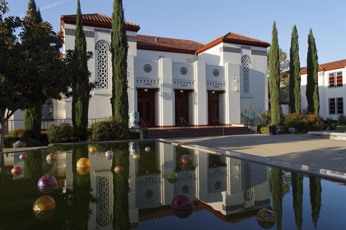 White Spanish Colonial Revival facade of the Campbell Heritage Theatre reflected in a courtyard pool with glass art spheres