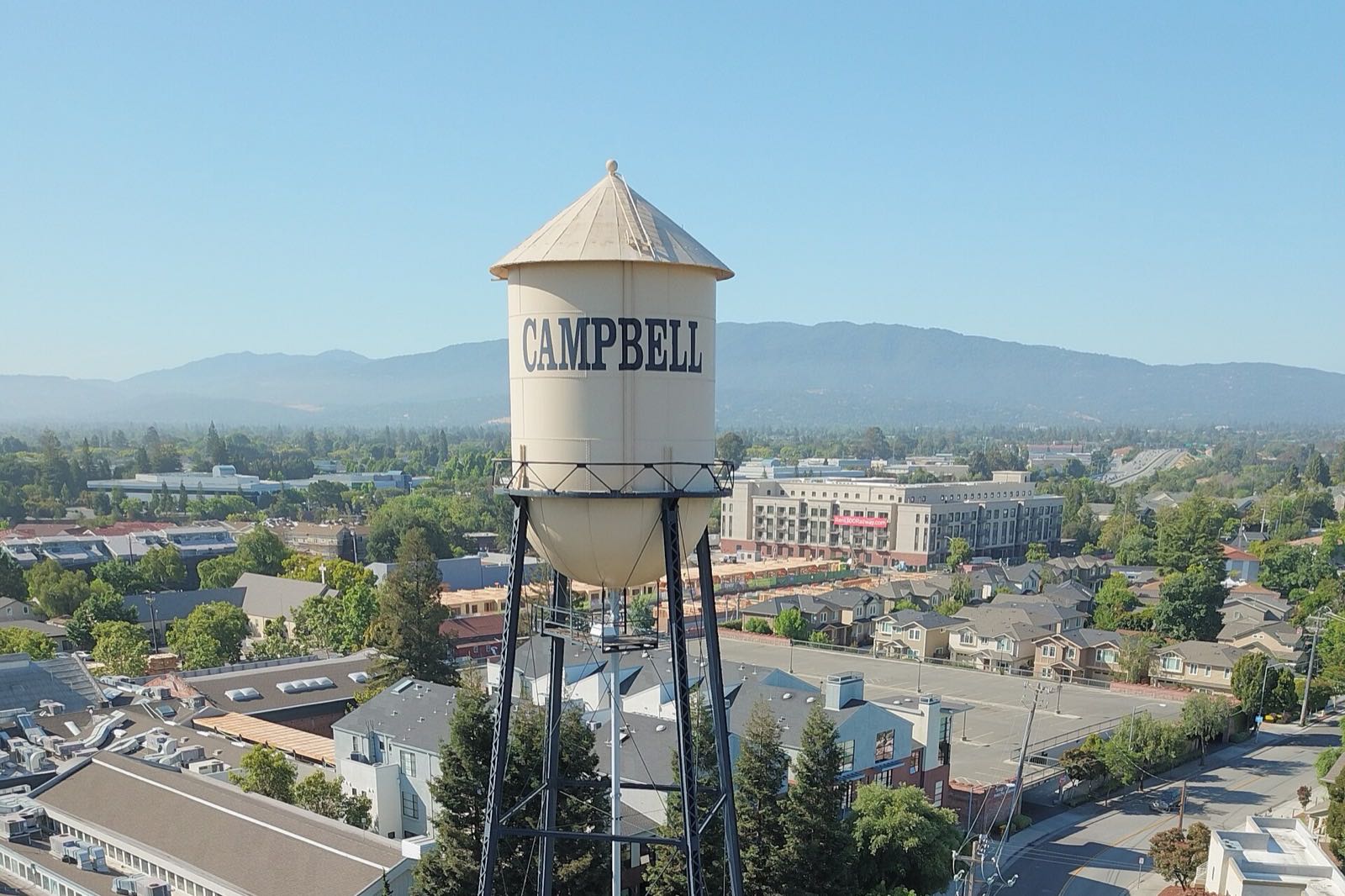 Aerial view of the cream-colored Campbell Water Tower with CAMPBELL painted on the tank, downtown rooftops below and the Santa Cruz Mountains behind on a clear day