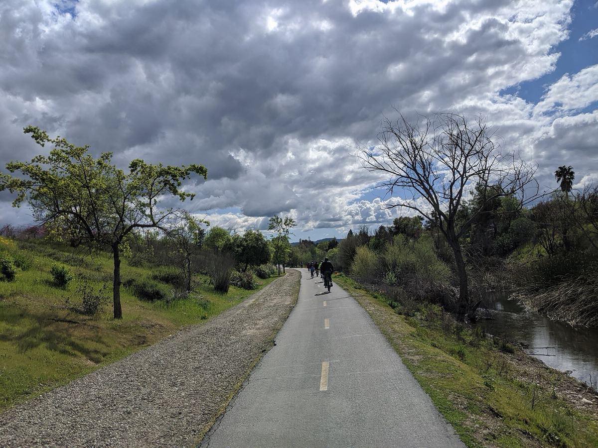 Cyclists riding the paved Los Gatos Creek Trail beside the creek in Campbell under big clouds