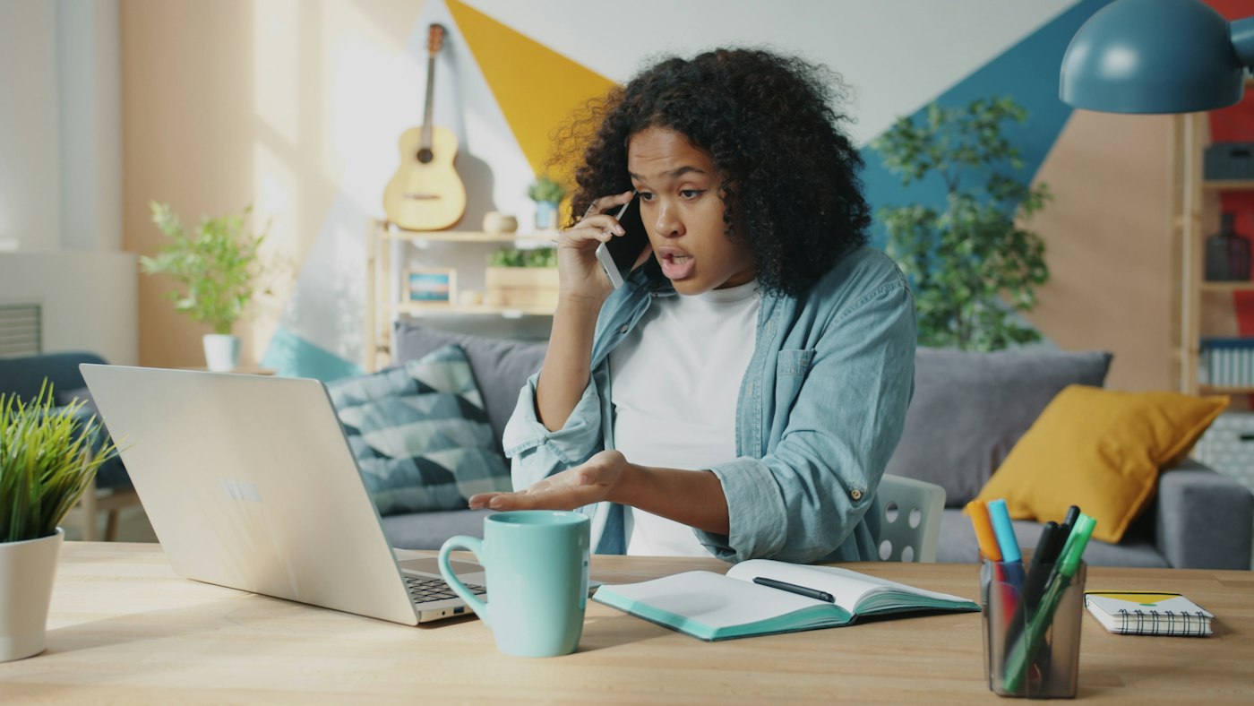 Young woman talking on her phone at a desk with a laptop, explaining a problem