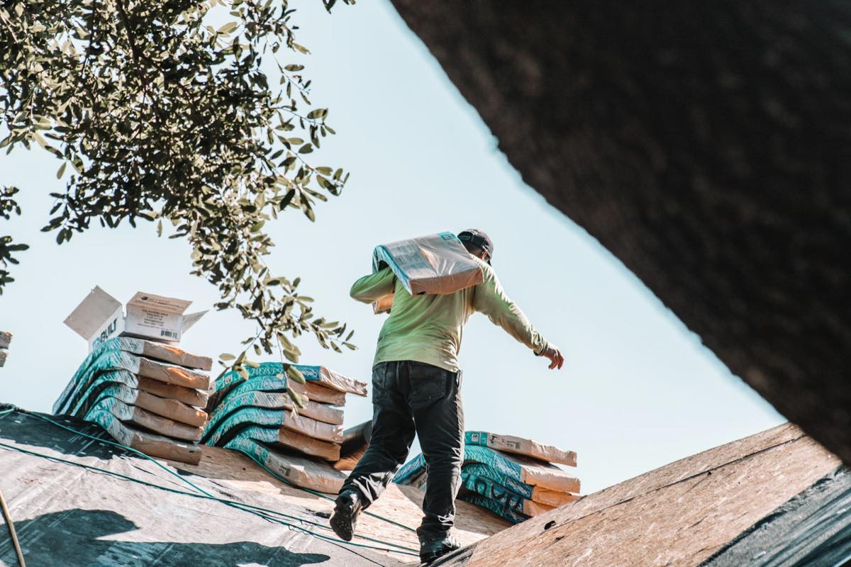 Roofer carrying a bundle of new shingles across a stripped roof with stacked shingle packs