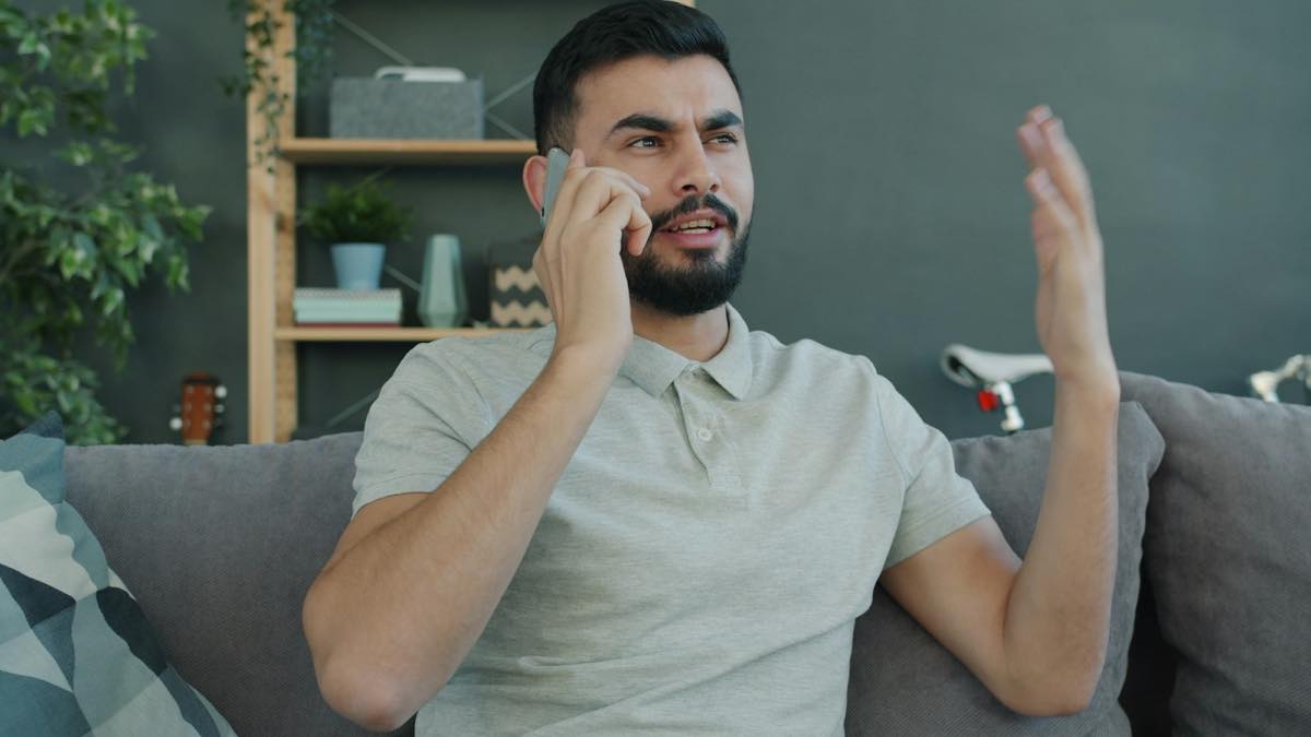 Man on a gray couch talking on his phone and gesturing with his other hand