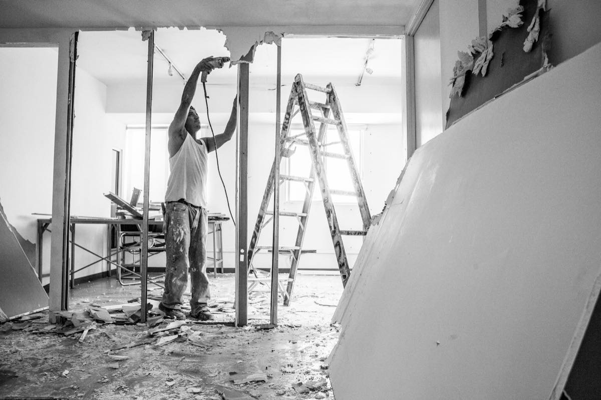 Worker in a stripped-out room cutting away ceiling drywall beside a ladder, with removed wallboard and debris on the floor