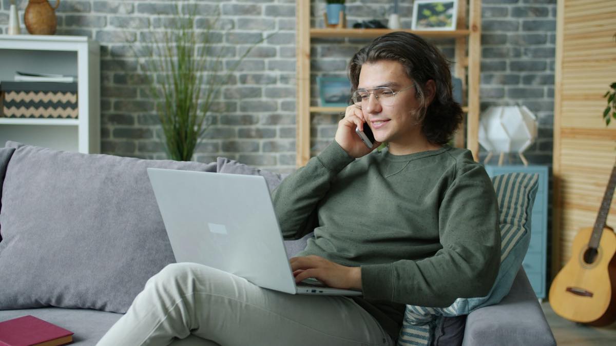 Young man in a green sweater on a phone call, laptop open on his lap, sitting on a grey sofa at home