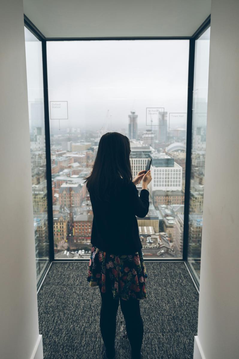 Woman seen from behind holding a smartphone as she stands at a tall window above a city