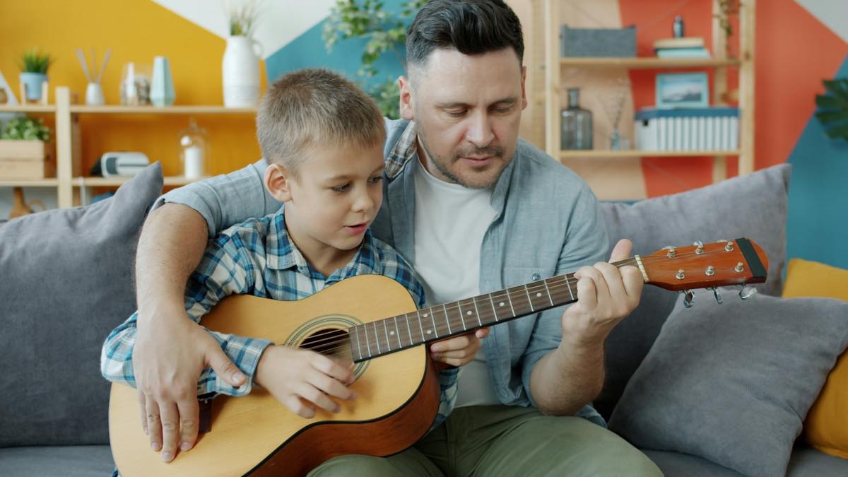 Father and son playing an acoustic guitar together on the living-room sofa