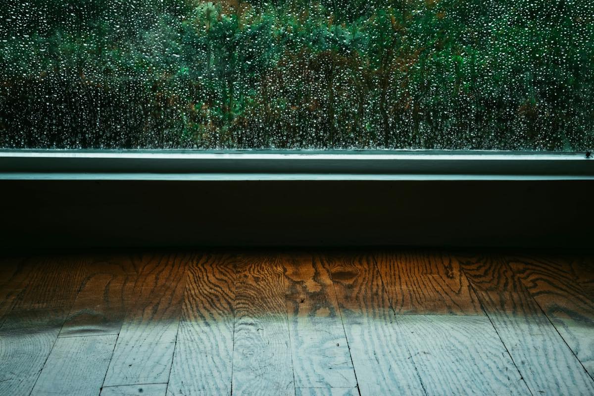Rain streaming down a window above a sill and a wide-plank wood floor