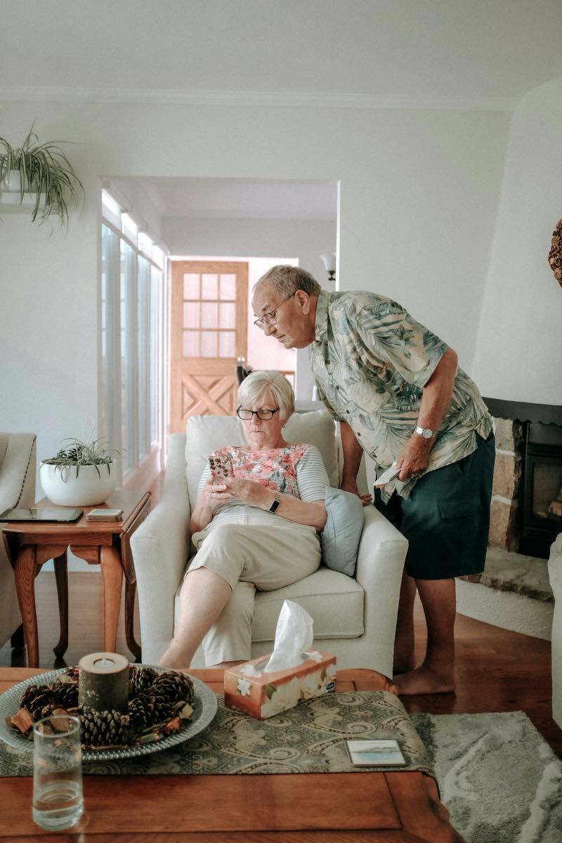 An older couple at home in a bright living room, the woman seated in an armchair looking at her phone while her husband leans in beside her