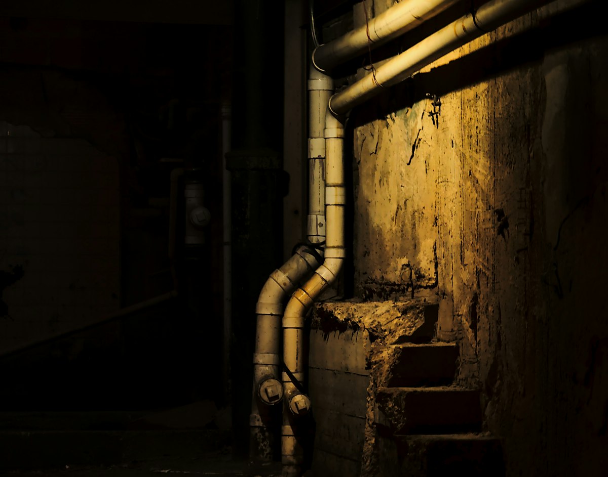 A dim utility room with exposed pipes below a window