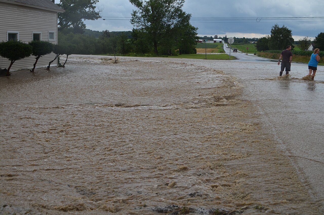 Brown floodwater sheeting across a lawn and a rural road beside a house, two people wading at the edge (Canandaigua, NY, July 2017)
