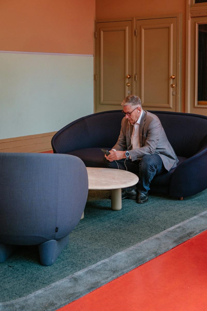 Gray-haired man in glasses and a gray blazer sitting on a navy sofa, looking down at his phone with earphones in