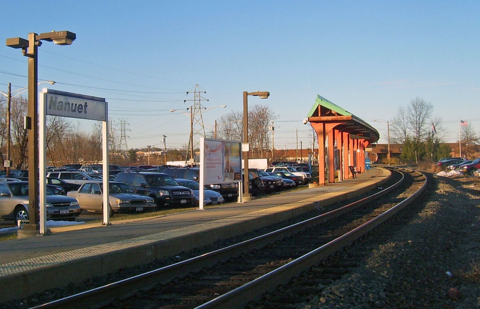Dark Nanuet station sign on the Pascack Valley Line platform, with the tracks, a red-posted shelter and the commuter lot under a clear sky