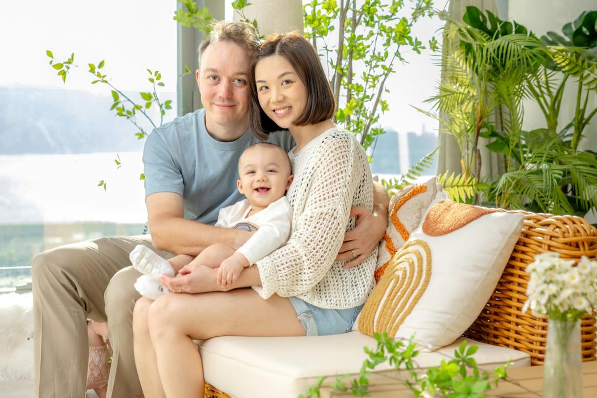 Smiling couple holding their laughing baby on a cushioned sofa in a bright room full of plants