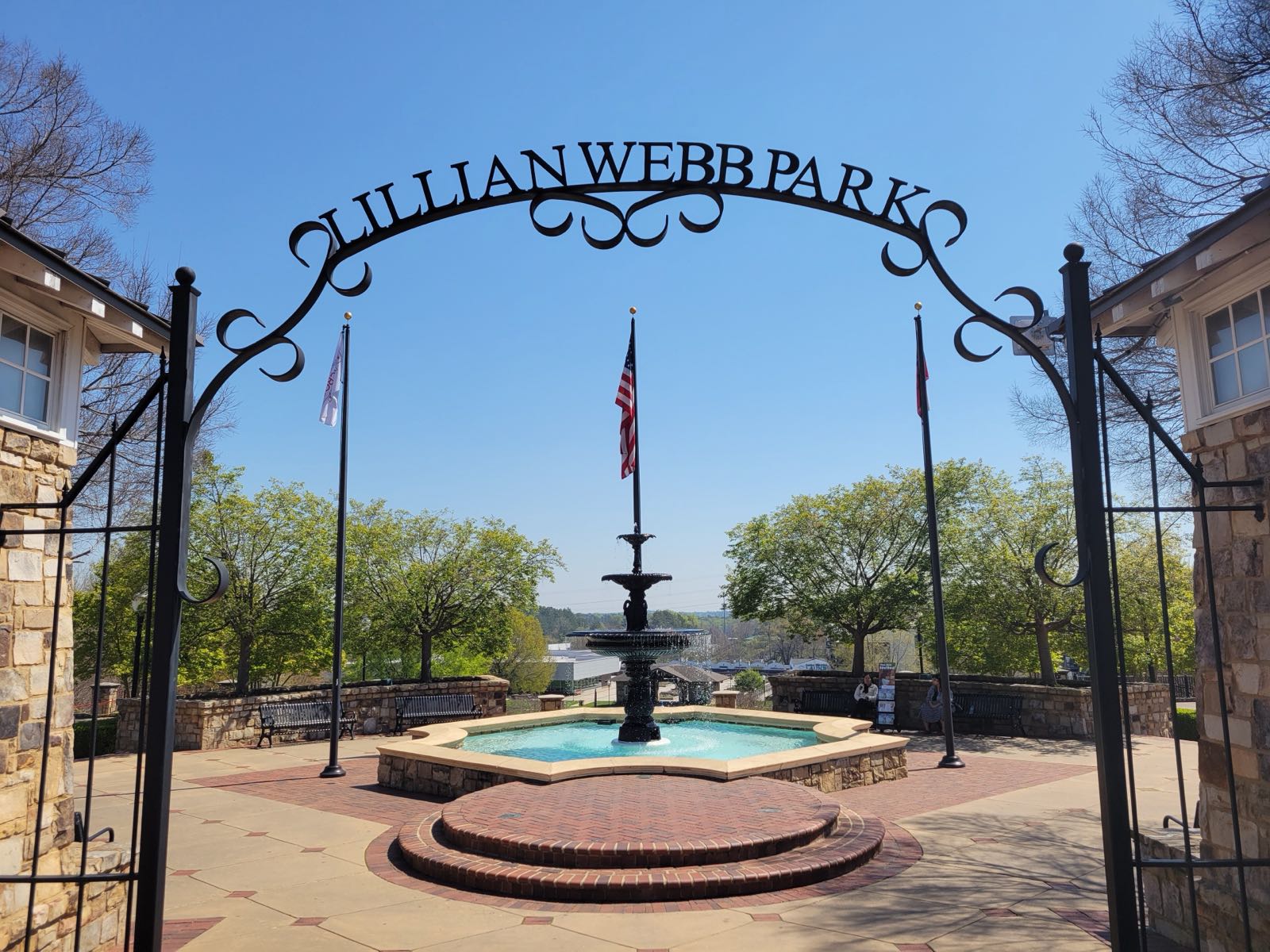 Wrought-iron Lillian Webb Park arch framing the tiered fountain and flags in downtown Norcross, GA, on a bright spring day