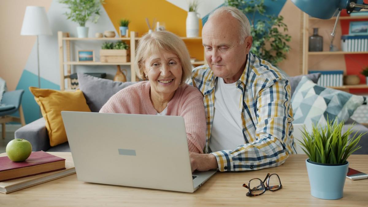 Older couple smiling together at a laptop on a table in their bright living room