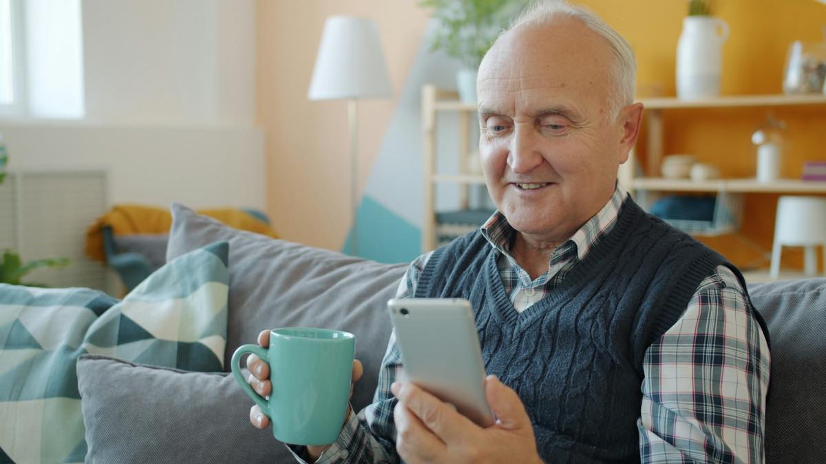 Smiling older man on a sofa at home, holding a mug and looking at his smartphone