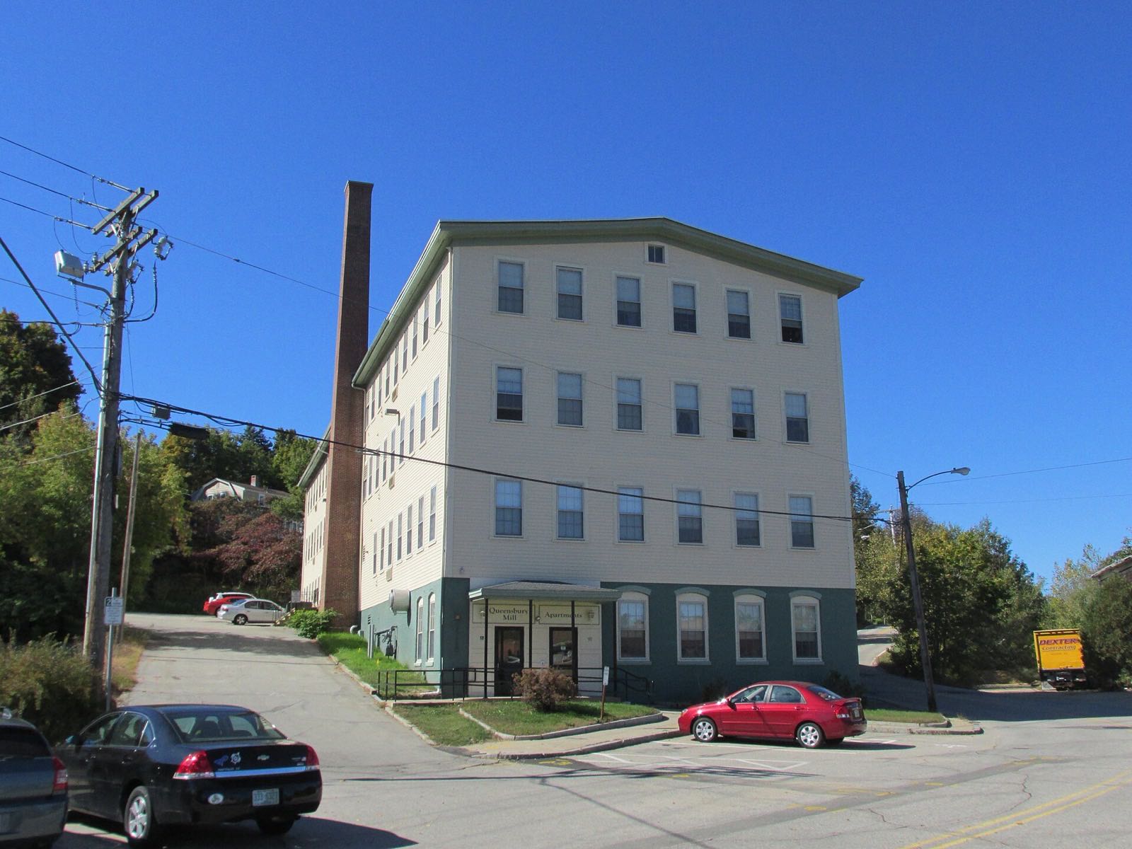 Queensbury Mill at 1 Market St, Somersworth: a four-story cream-painted former textile mill beside its brick smokestack, under a clear blue sky
