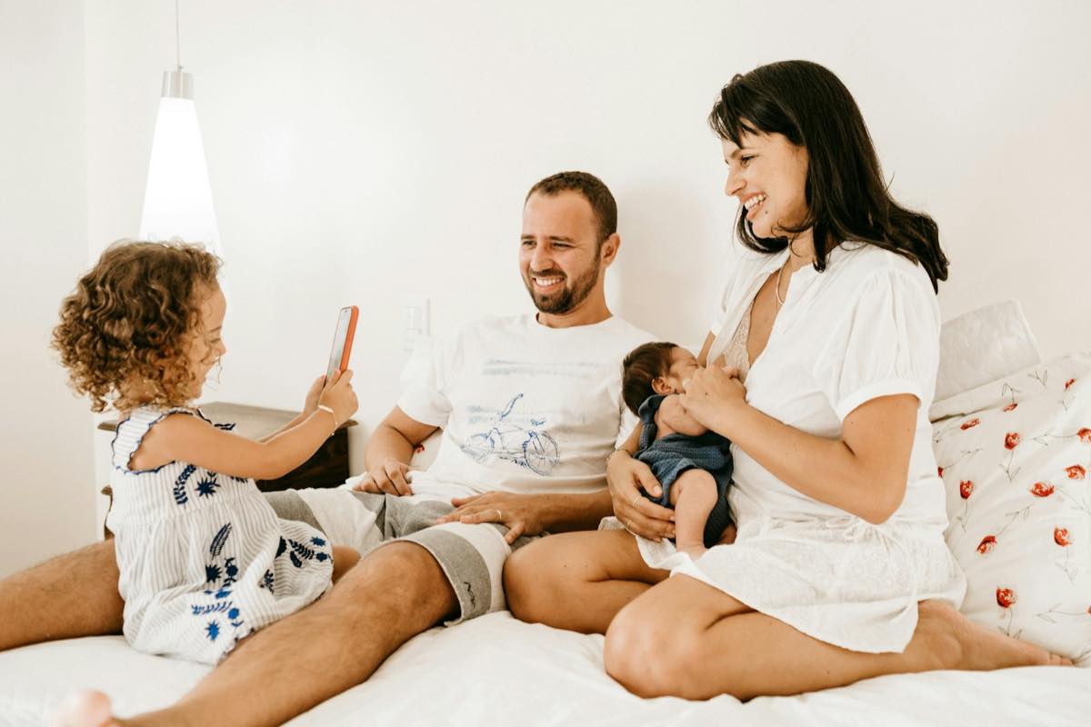 Smiling parents sitting on a bed holding their newborn while their curly-haired toddler holds up a phone